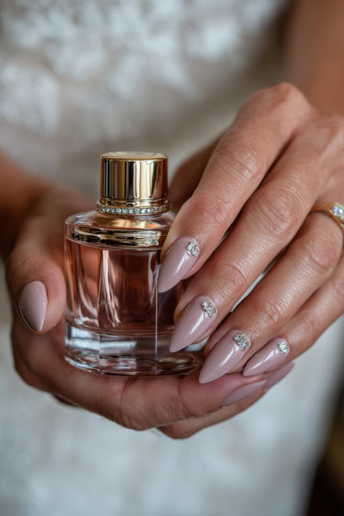 Bride holding a rose gold perfume bottle, almond nails in pastel mauve with shimmer overlay and crystal-studded accents on the ring fingers 