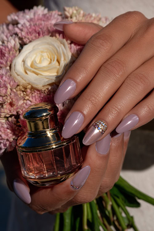 Bride holding a rose gold perfume bottle, almond nails in pastel mauve with shimmer overlay and crystal-studded accents on the ring fingers