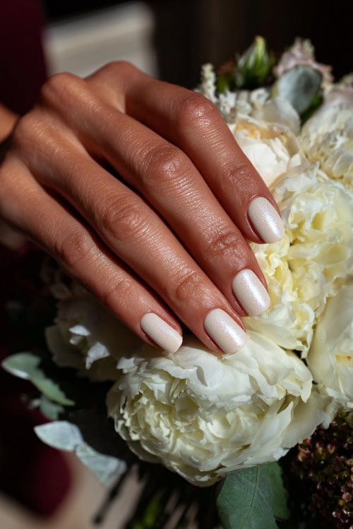 Bride’s hand beside floral decor, square-shaped nails in creamy ivory with shimmer topcoat and tiny pearl accents along the base of each nail 