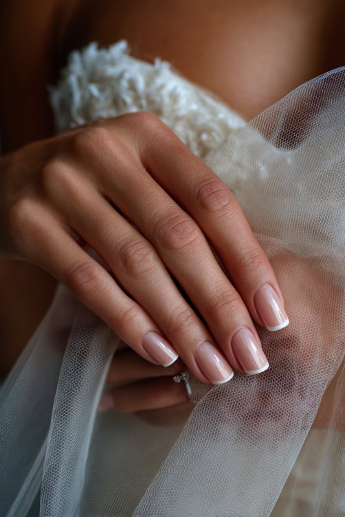 Bride’s hand gently brushing tulle gown, short square nails in creamy beige with chrome French tips and tiny crystal accents at the cuticle 