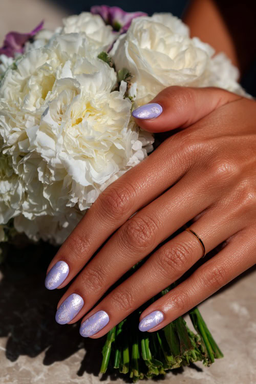 Bride’s hand resting beside wedding bouquet, medium almond nails in soft pastel lavender with a matte finish and white floral stamping