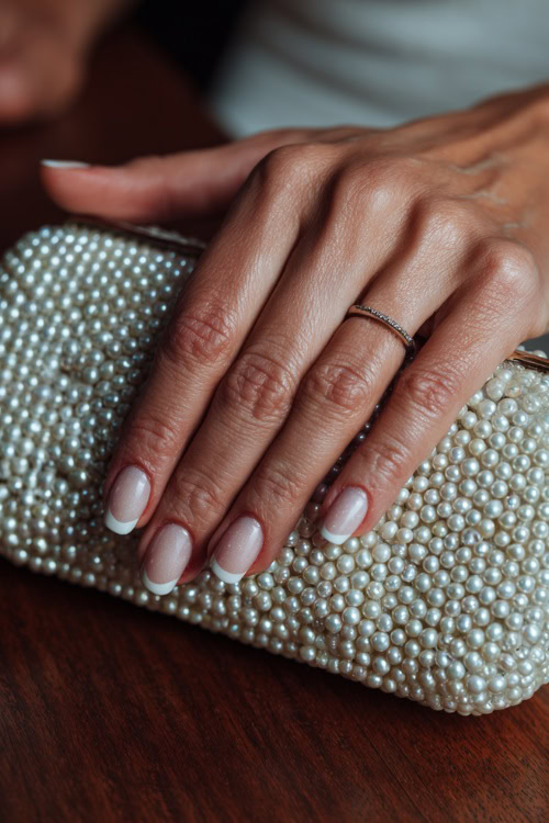 Bride’s hand resting on a pearl clutch, short square nails in sheer ivory with classic French tips and subtle shimmer