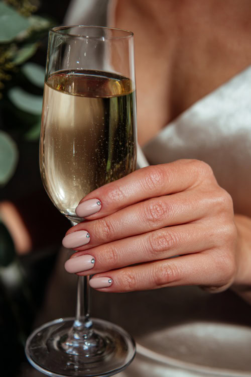 Bridesmaid’s hand holding a champagne glass, short ballerina nails in pastel beige with shimmer overlay and small crystal accents near the tips 