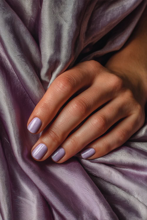 Bridesmaid’s hand resting on satin dress fabric, short square nails in pastel lavender with shimmer overlay and silver dot details at the base of each nail 
