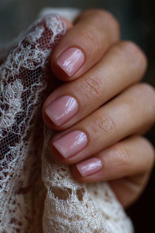 Close-up of a hand holding a lace-trimmed veil, short square nails in soft blush pink with classic French tips and a glossy finish 