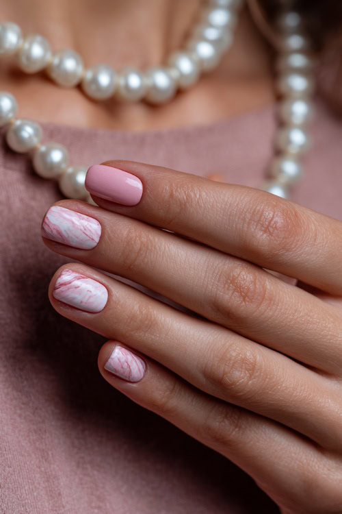 Close-up of hand brushing pearl necklace, short square nails in pale pink with satin finish and one accent nail in marble swirl design 
