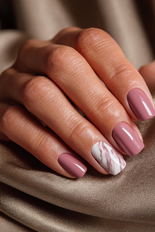 Close-up of hand resting on silk fabric, medium square nails in dusty rose with glossy finish and one accent nail in white marble swirl design 