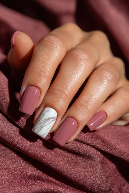 Close-up of hand resting on silk fabric, medium square nails in dusty rose with glossy finish and one accent nail in white marble swirl design 