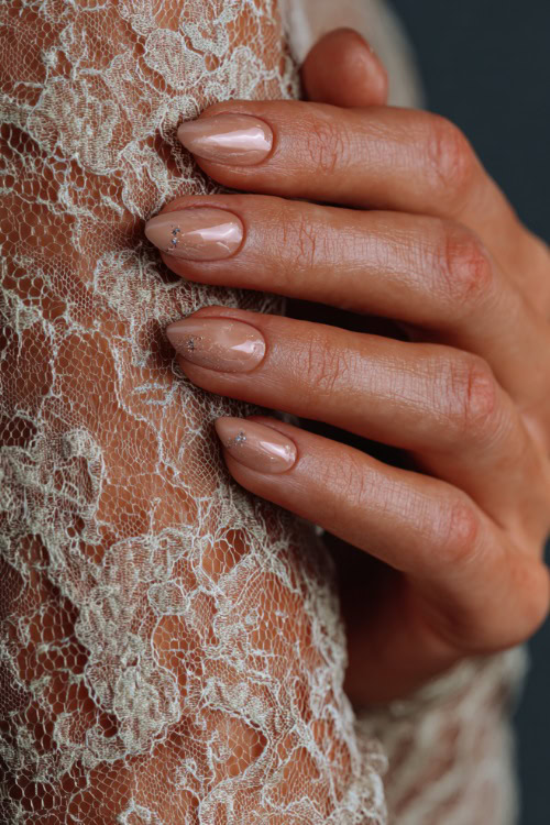 Elegant hand adjusting lace dress sleeve, almond nails in soft beige with glossy finish and tiny crystal-studded cuticle details on each finger 