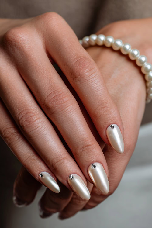 Hand adjusting a pearl bracelet, almond-shaped nails in creamy ivory with silver chrome finish and dainty rhinestones at the cuticle