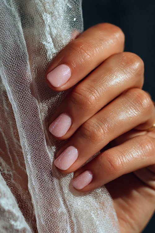 Hand gently lifting bridal veil, short square nails in pastel pink with fine silver shimmer and tiny crystal accents on the middle fingers