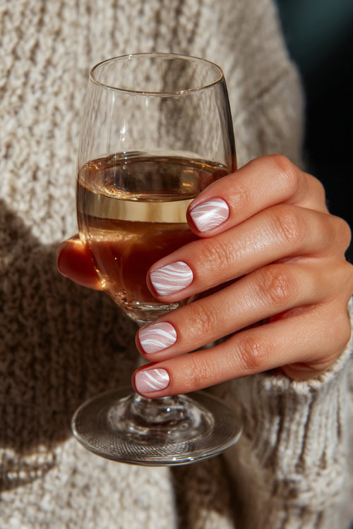 Hand holding a glass of champagne, short square nails in soft beige-pink with shimmer overlay and one accent nail with minimal white swirl design 