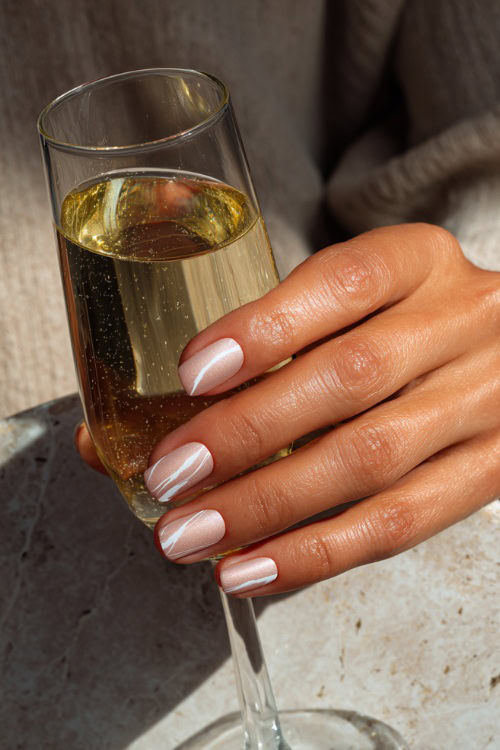 Hand holding a glass of champagne, short square nails in soft beige-pink with shimmer overlay and one accent nail with minimal white swirl design
