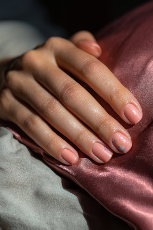 Hand resting beside satin pillow, medium square nails in pastel peach with chrome shimmer and one floral accent on the ring finger 