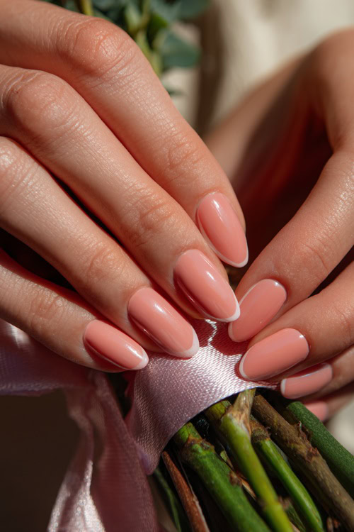 Hand tying a ribbon on a bouquet, ballerina-shaped nails in pale peach with chrome French tips and a glossy finish