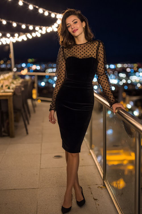 Fashion-forward woman in a black velvet wedding guest dress with sheer polka-dot long sleeves and pointed heels, standing at an evening city rooftop wedding with bistro lights above 