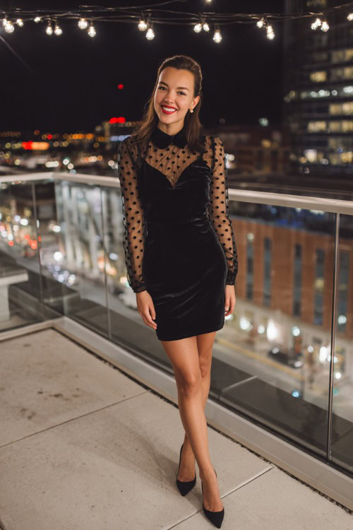 Fashion-forward woman in a black velvet wedding guest dress with sheer polka-dot long sleeves and pointed heels, standing at an evening city rooftop wedding with bistro lights above