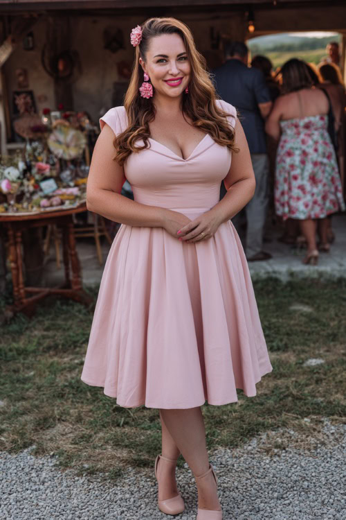 Full body of a plus-size woman in a pastel pink fit-and-flare wedding guest dress with cap sleeves, floral earrings, and block heels, standing near a countryside wedding with vintage decor