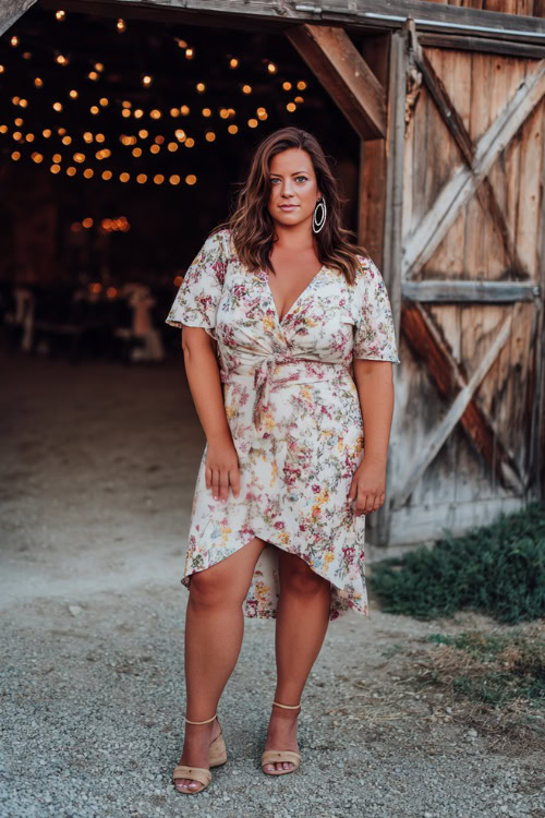 Full body of a plus-size woman in a short-sleeve floral high-low wedding guest dress with a tie waist, wedge heels, and hoop earrings 