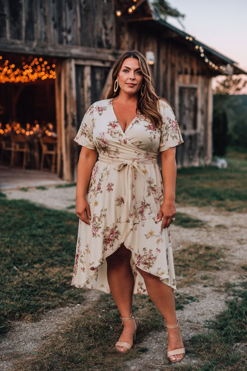 Full body of a plus-size woman in a short-sleeve floral high-low wedding guest dress with a tie waist, wedge heels, and hoop earrings