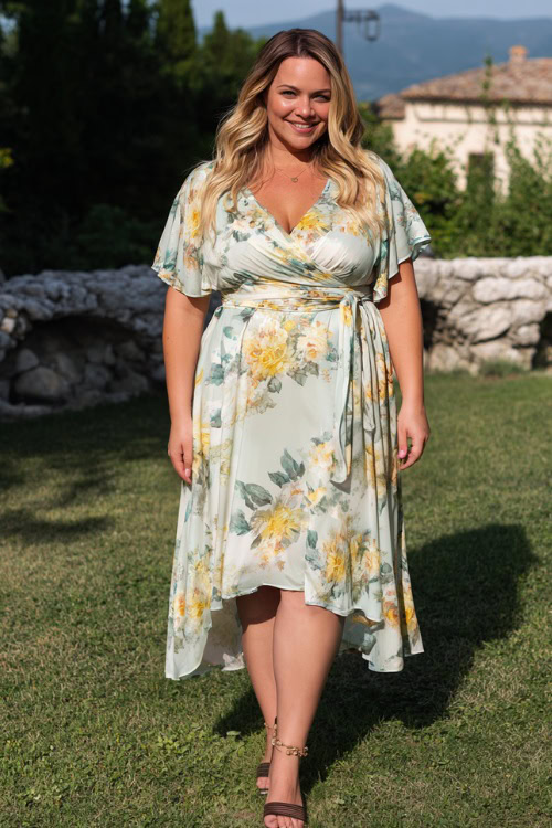 Full body of a plus-size woman wearing a flowy floral wrap wedding guest dress with flutter sleeves and a midi hemline, paired with ankle-strap heels