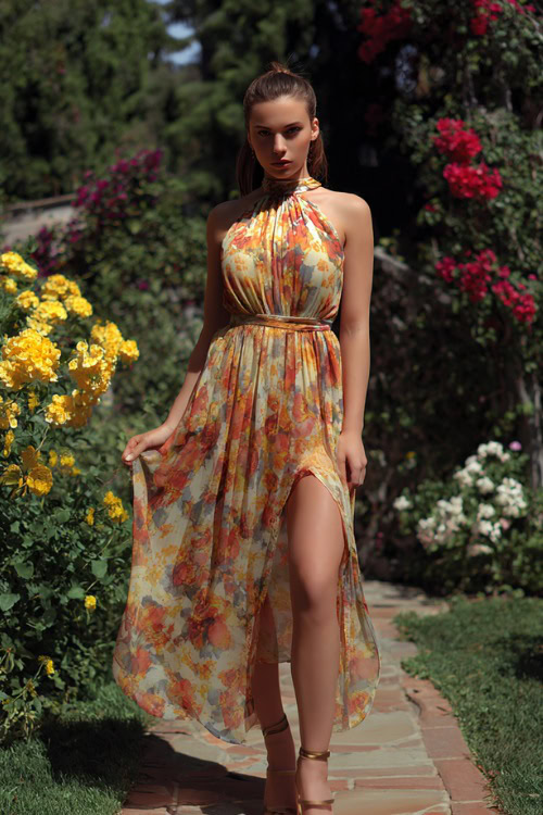 Full body of a young woman wearing a sleeveless floral chiffon wedding guest dress with a flowy skirt and strappy heels, standing in a sunny garden with blooming flowers