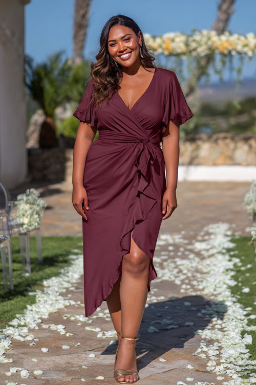 Plus-size woman wearing a burgundy midi wedding guest dress with flutter sleeves and ruching at the waist, paired with gold strappy sandals, posing near a decorated aisle with white petals