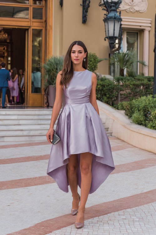 Stylish woman in a sleeveless high-low wedding guest dress with a satin finish, soft lavender color, carrying a clutch bag, standing at an elegant hotel entrance