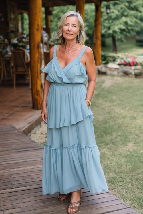 Stylish woman over 50 wearing a flowy A-line wedding guest dress with soft ruffles, sleeveless, sky blue color, layered skirt, wearing metallic sandals 