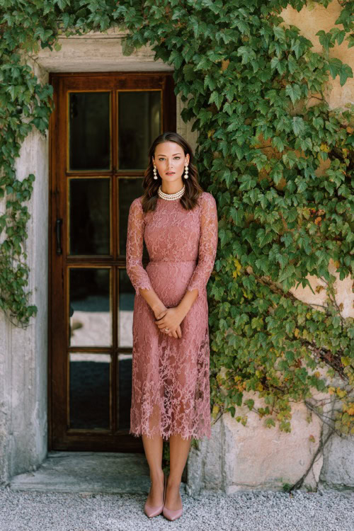 Woman in a dusty rose lace midi dress with sheer long sleeves and a scalloped neckline, paired with pearl jewelry and classic pumps, standing beside an ivy-covered wall at a vintage wedding venue