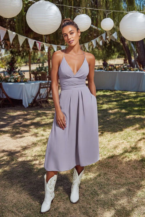 Woman in a sleek V-neck midi dress in soft lavender paired with white stitched cowboy boots, posing under hanging lanterns and bunting at a backyard wedding celebration 