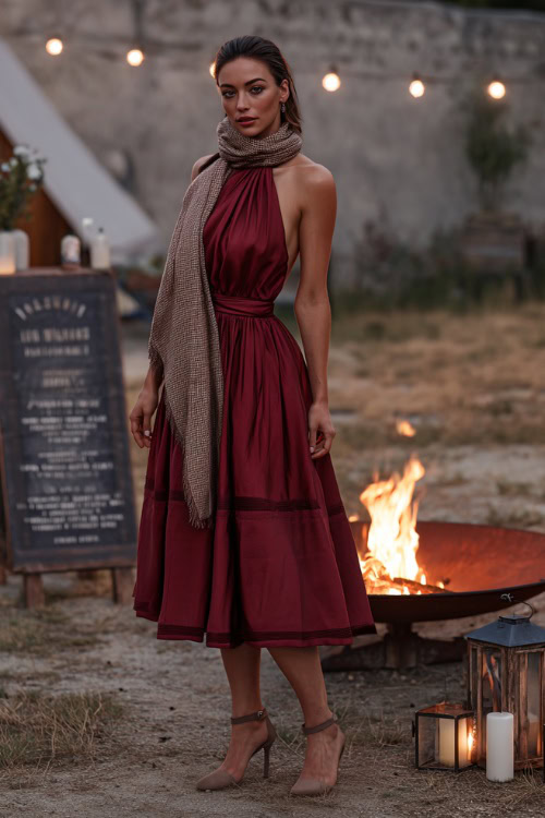 Woman in a sleeveless dark red midi dress with a high neckline and layered skirt, styled with a shawl and heeled mules, posing beside a fire pit and wedding signage