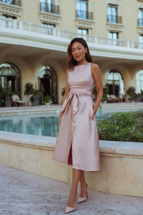 Woman in a sleeveless satin midi wedding guest dress with a bow detail at the waist, pale blush color, wearing classic heels