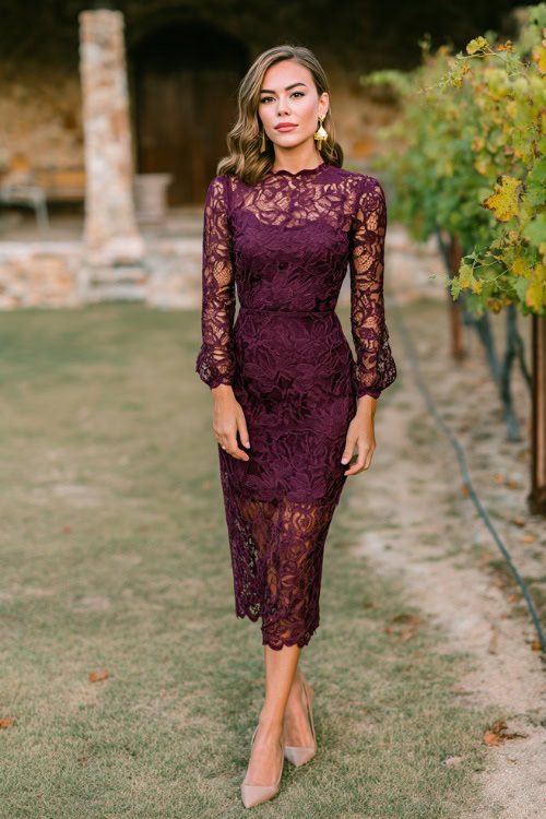 Woman wearing a deep plum lace midi dress with scalloped hem and long sleeves, styled with gold earrings and taupe heels, standing at a winery wedding in autumn 