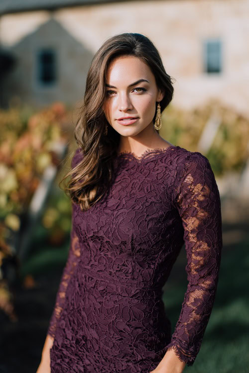 Woman wearing a deep plum lace midi dress with scalloped hem and long sleeves, styled with gold earrings and taupe heels, standing at a winery wedding in autumn