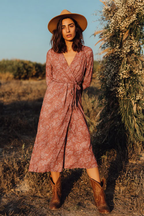 Woman wearing a dusty rose wrap midi wedding guest dress with a tie waist, styled with classic brown leather cowboy boots and a wide-brim hat 