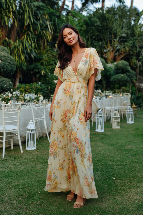 Woman wearing a long floral chiffon maxi dress with short flutter sleeves, platform sandals, and subtle jewelry, posing in a lush garden wedding setup with white chairs and lanterns 